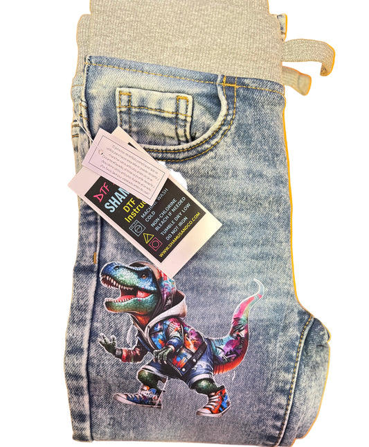 Denim shorts with a colorful dinosaur design and a tag on a white background Customized toddler jeans featuring a colorful dinosaur DTF design on the pant leg