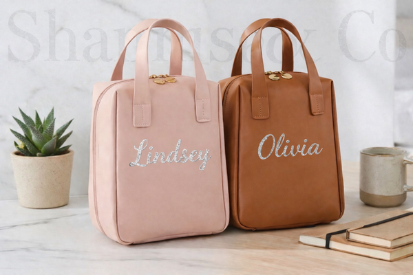 Two handbags, one pink and one brown, with names embroidered on them, placed on a surface with a plant and mug in the background.