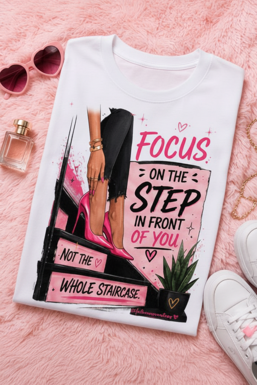 White t-shirt with motivational graphic and text on a pink surface with shoes and sunglasses.