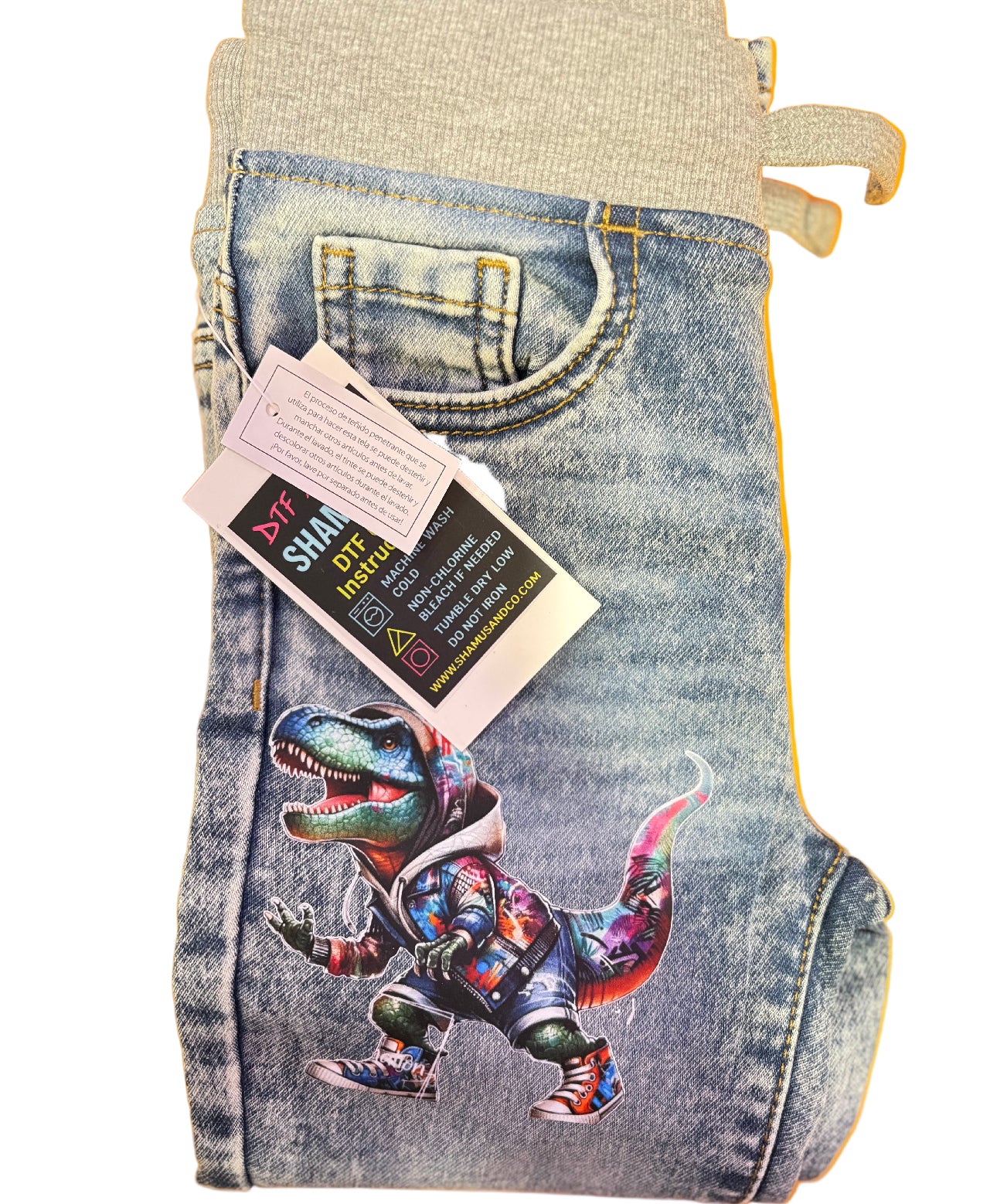 Denim shorts with a colorful dinosaur design and a tag on a white background Customized toddler jeans featuring a colorful dinosaur DTF design on the pant leg