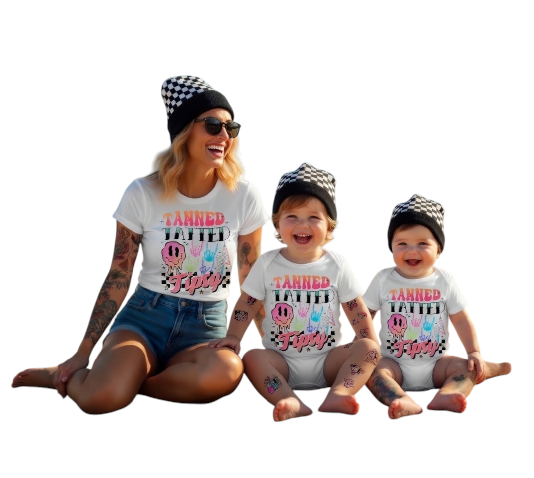 Woman and two children sitting on a beach wearing matching 'Tanned Tatted Tipsy' shirts with product tags displayed.