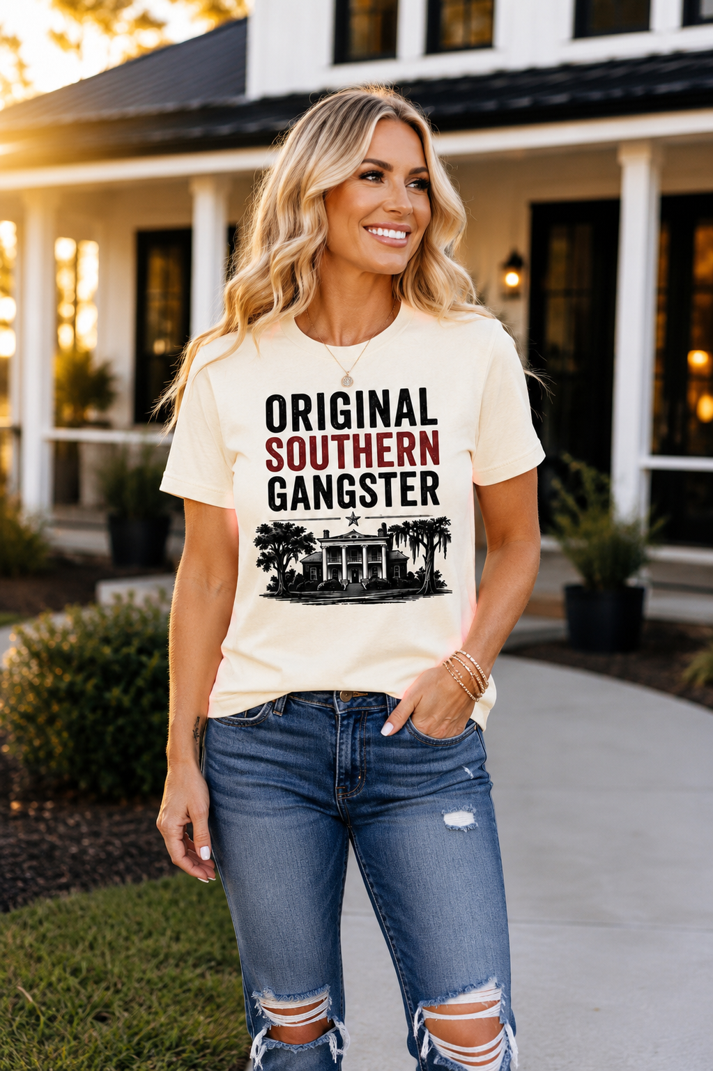 A woman wearing a cream-colored graphic t-shirt with the text 'ORIGINAL SOUTHERN GANGSTER' in black and red, paired with distressed blue jeans, standing in front of a house.