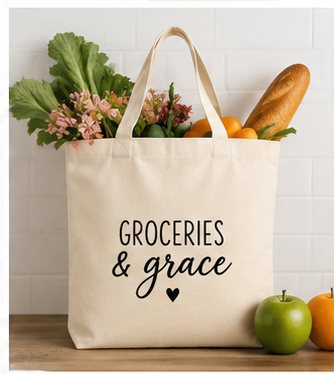 A cream canvas tote bag with black text reading 'GROCERIES & grace' and a small heart, filled with groceries including a baguette, greens, flowers, and oranges, set against a white subway tile wall.