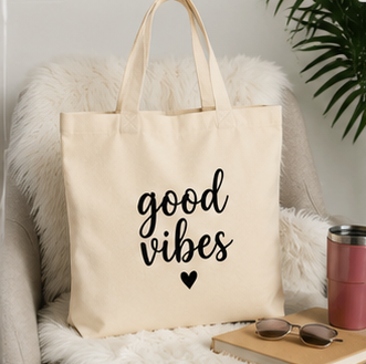 A cream canvas tote bag with black script text reading 'good vibes' and a small heart, displayed on a white faux-fur chair in a styled indoor setting with sunglasses, a pink tumbler, a book, and a plant.