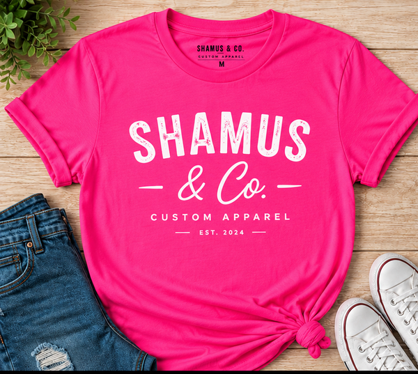 Pink t-shirt with 'Shamus & Co. Custom Apparel' text on a wooden surface with jeans and shoes.