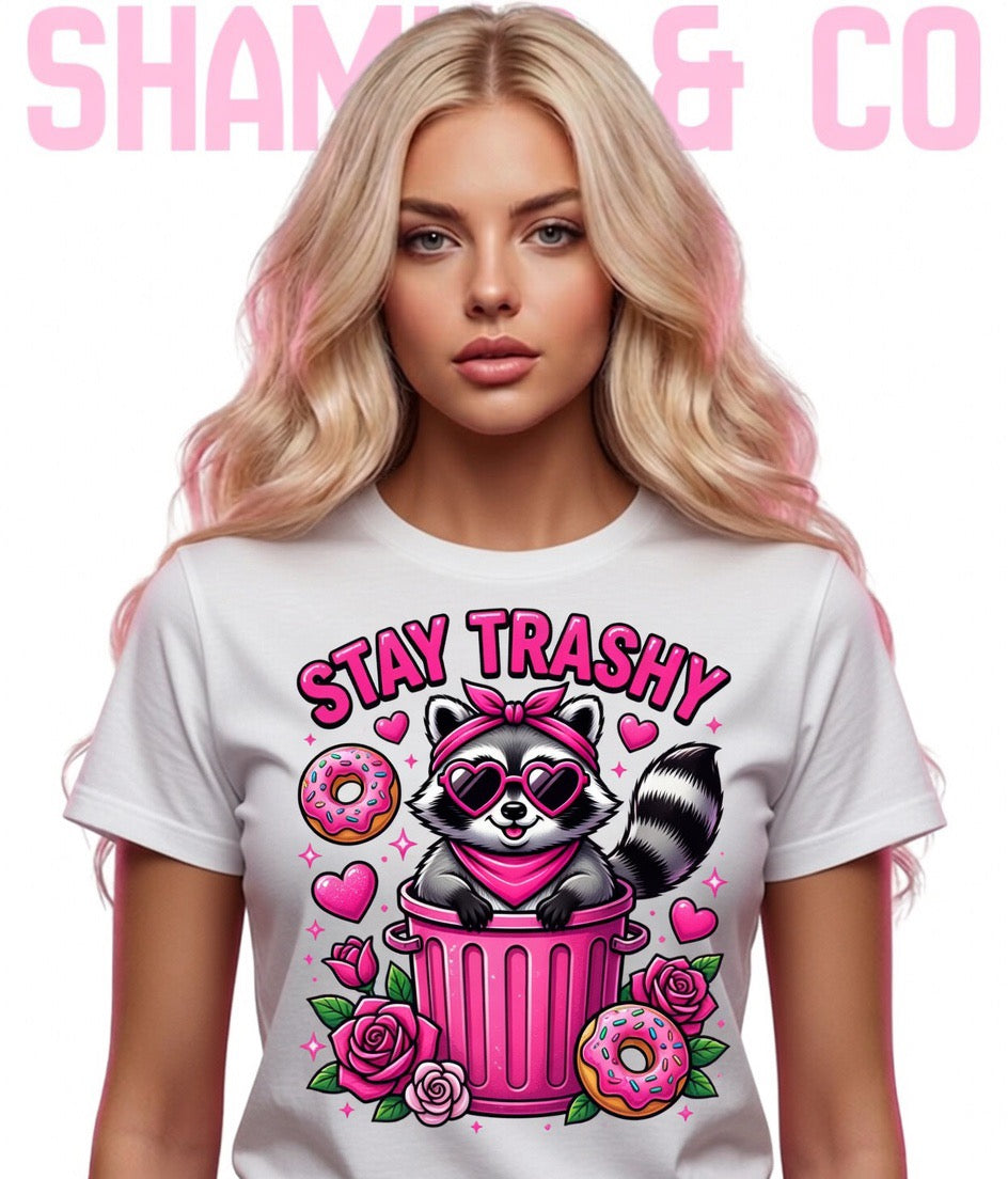 Woman wearing a white t-shirt with a graphic design featuring a raccoon, pink trash can, and text 'Stay Trashy'. Brand 'shamus & Co' is visible.