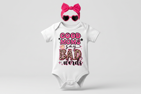 Baby onesie with 'Good moms say bad words' text, pink bow, and heart-shaped sunglasses on a light gray background