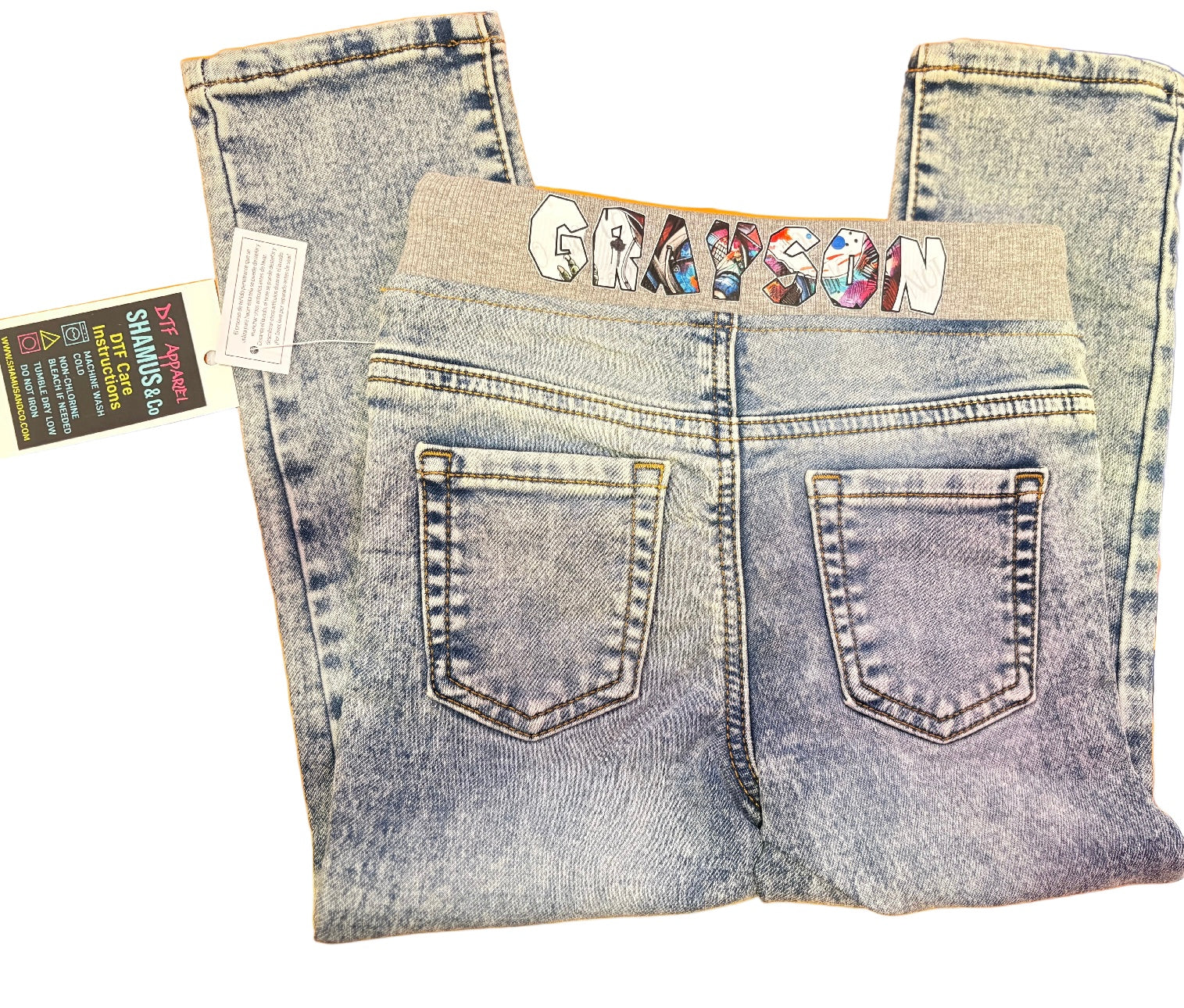 Folded toddler jeans with personalized name ‘GRAY’ on a gray waistband, displayed on an orange background.