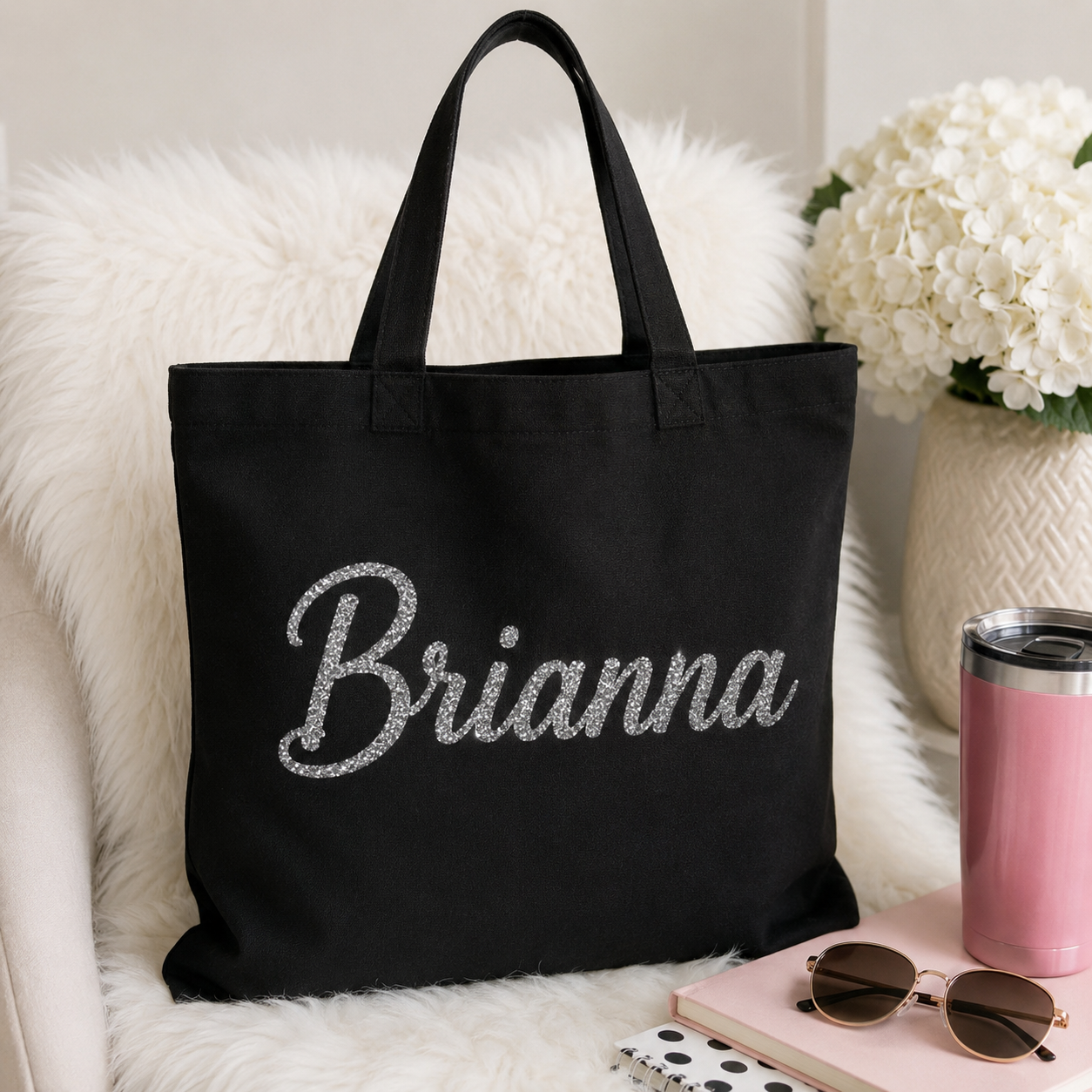 Personalized black canvas tote bag with a custom silver glitter HTV name, displayed with a pink tumbler, sunglasses, notebook, and white flowers in a stylish lifestyle setting.