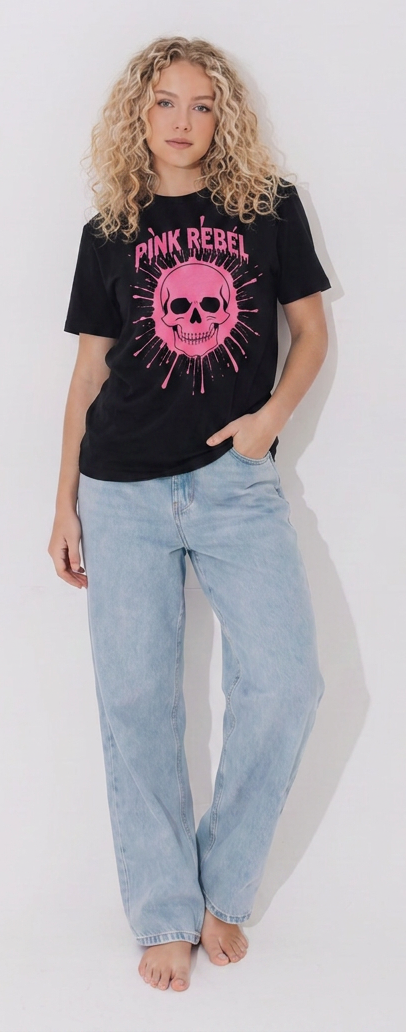 A blonde woman wearing a black Pink Rebel graphic streetwear t-shirt with a bright pink dripping skull design, paired with light blue baggy jeans, standing against a clean white wall.